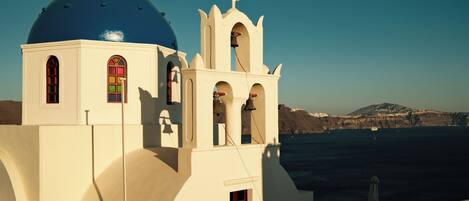 A white building with a blue dome and a bell tower, overlooking a body of water and mountains.