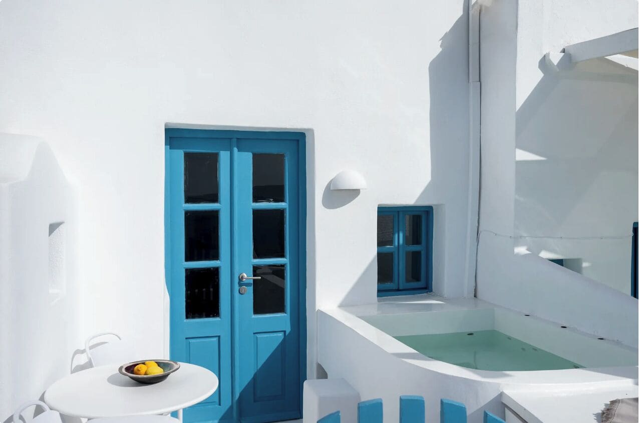 A white building with a blue door and window, a small table with a bowl of fruit, and a white wall with a light fixture.