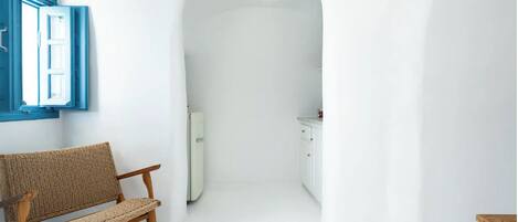 Un pasillo estrecho con un banco de madera a la izquierda, una puerta blanca al final y una pequeña zona de cocina con armarios y una encimera.