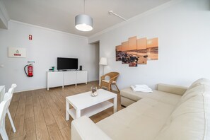 A modern living room with a white sofa, a white coffee table, a television on a white cabinet, a wicker chair, and a wall-mounted artwork. - Apartment 'T1 Superior' with Shared Pool, Wi-Fi and Air Conditioning (Portimão)
