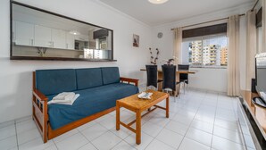 A living room with a sofa, a small wooden table, a mirror, a dining area with chairs, and a view of buildings through a window. - Apartment 'T2 Premium' with Shared Pool, Wi-Fi and Air Conditioning (Portimão)
