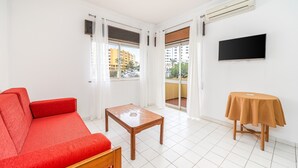 Ett rum med en röd soffa, ett träbord, en tv på väggen och en balkong med utsikt över byggnader.
