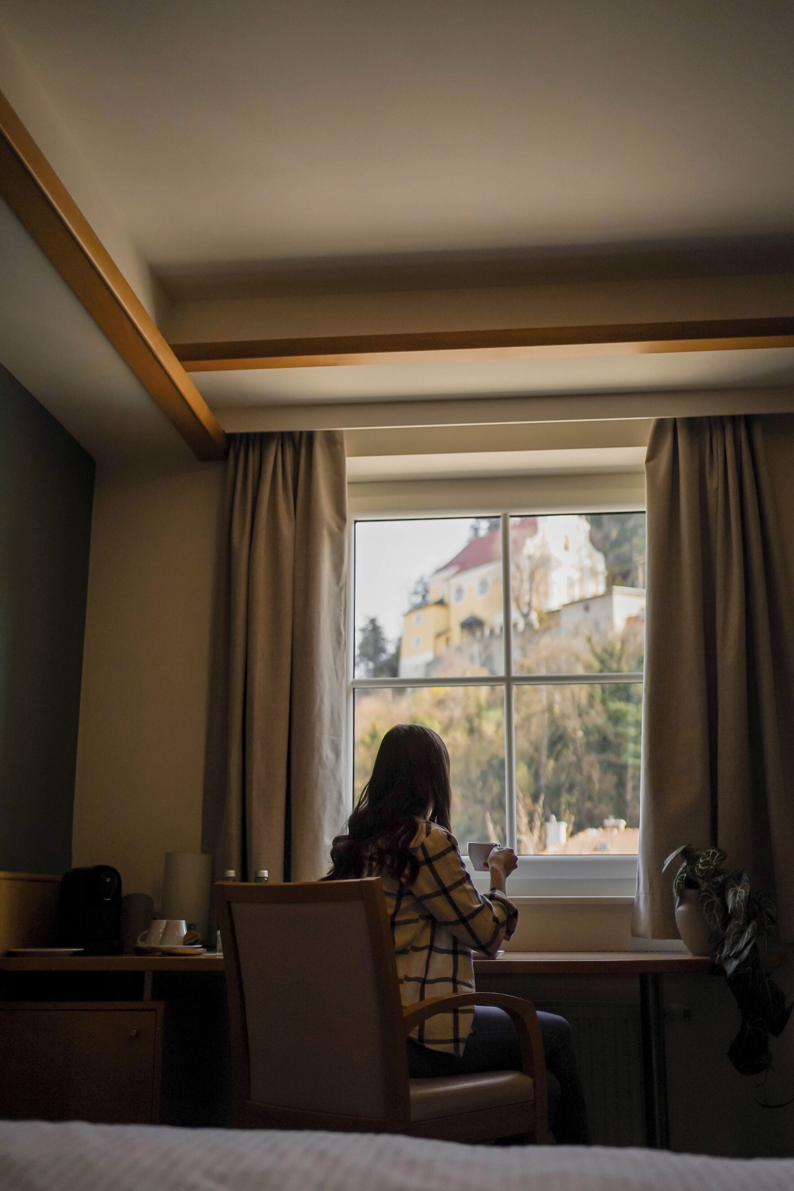 A room with a bed, a desk with a chair, a window with curtains, and a view of buildings outside.