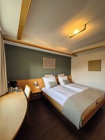 Quarto de hotel com cama, mesas de cabeceira, uma cadeira e luminária fixa na parede.