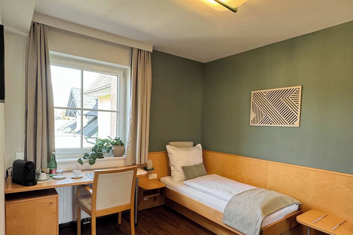 Une pièce de taille modeste comprenant un lit, un bureau, une chaise, une fenêtre avec des rideaux, un luminaire fixé au mur et une plante en pot.