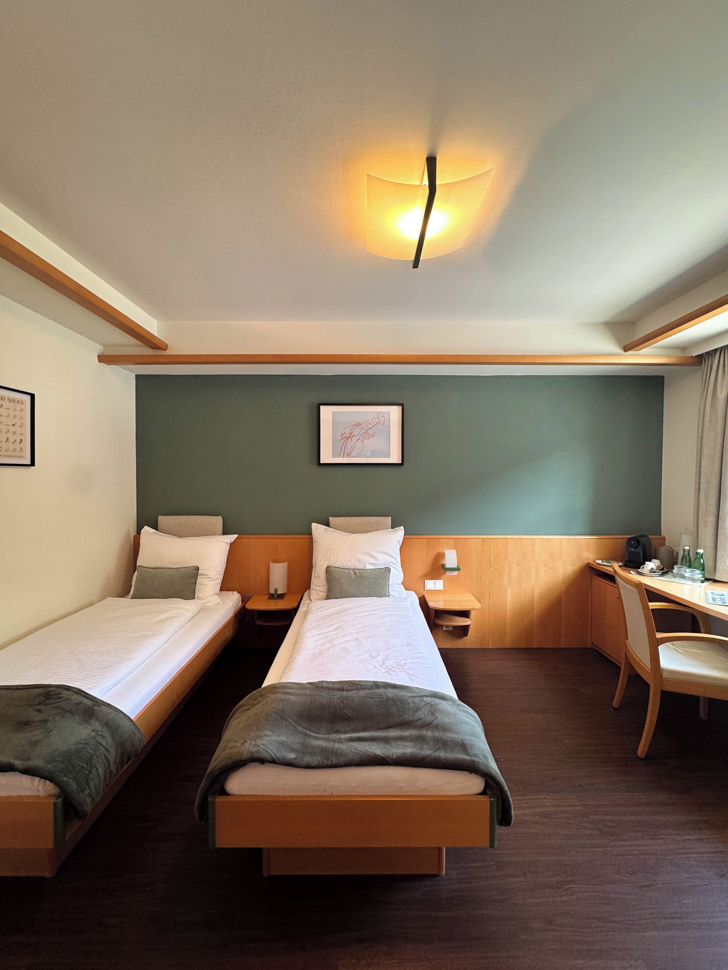 Une chambre d’hôtel avec deux lits simples, un bureau, une chaise, une table de chevet avec une lampe et un tableau encadré au mur.
