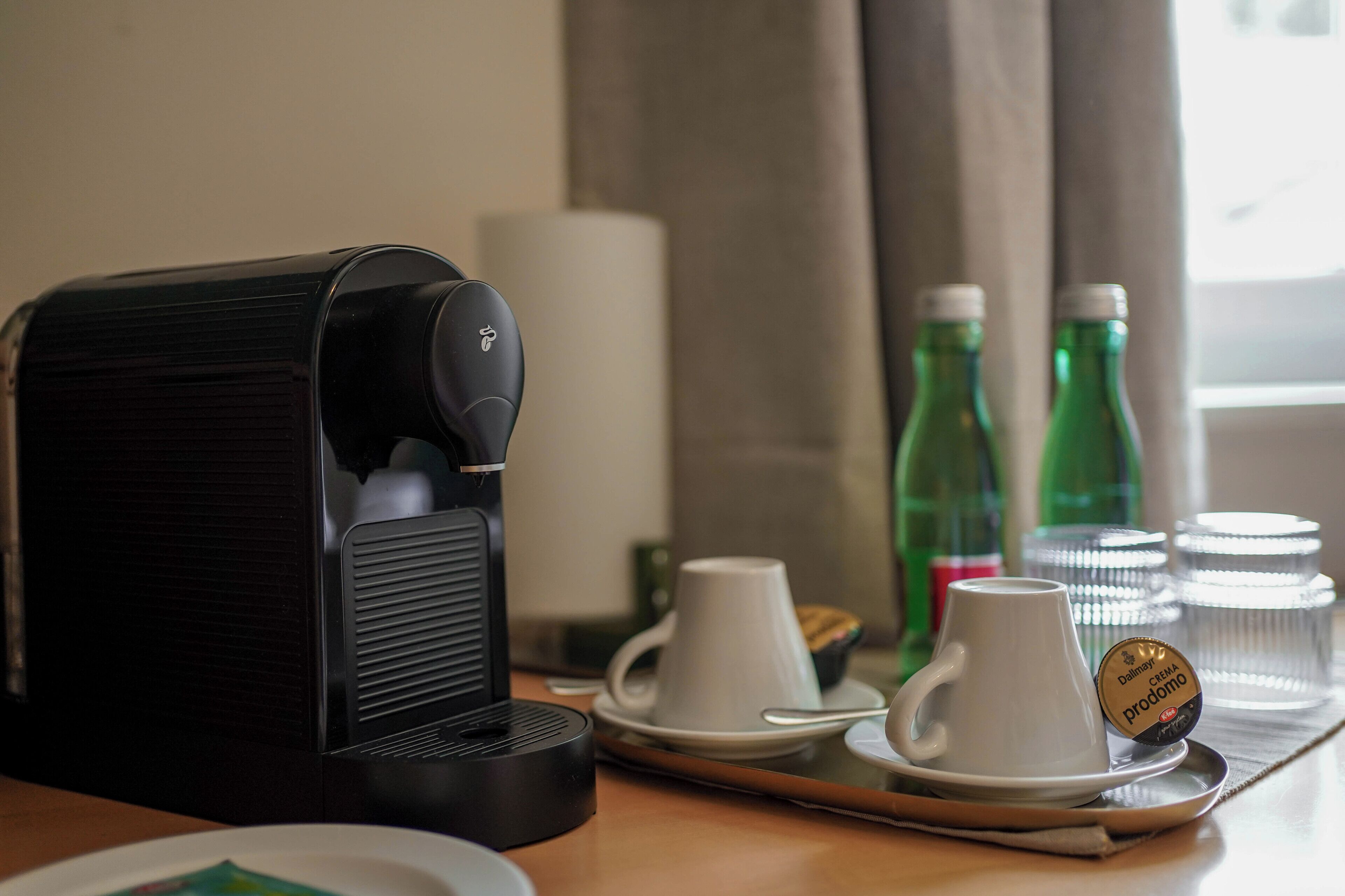 Kaffemaskine, to hvide kopper, et fad og grønne flasker på et bord.