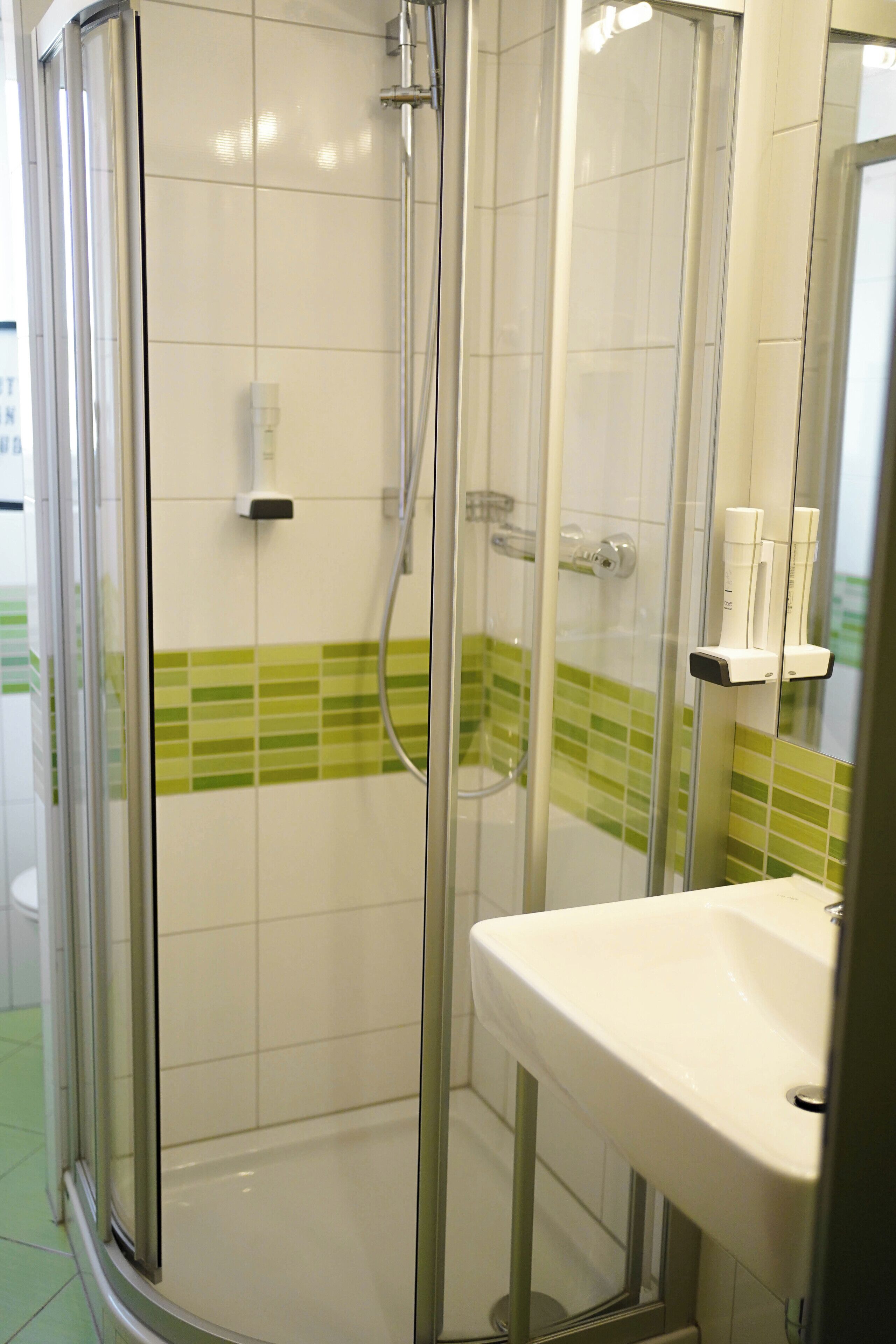 Una ducha con cerramiento de vidrio, un lavabo blanco y paredes con azulejos verdes.