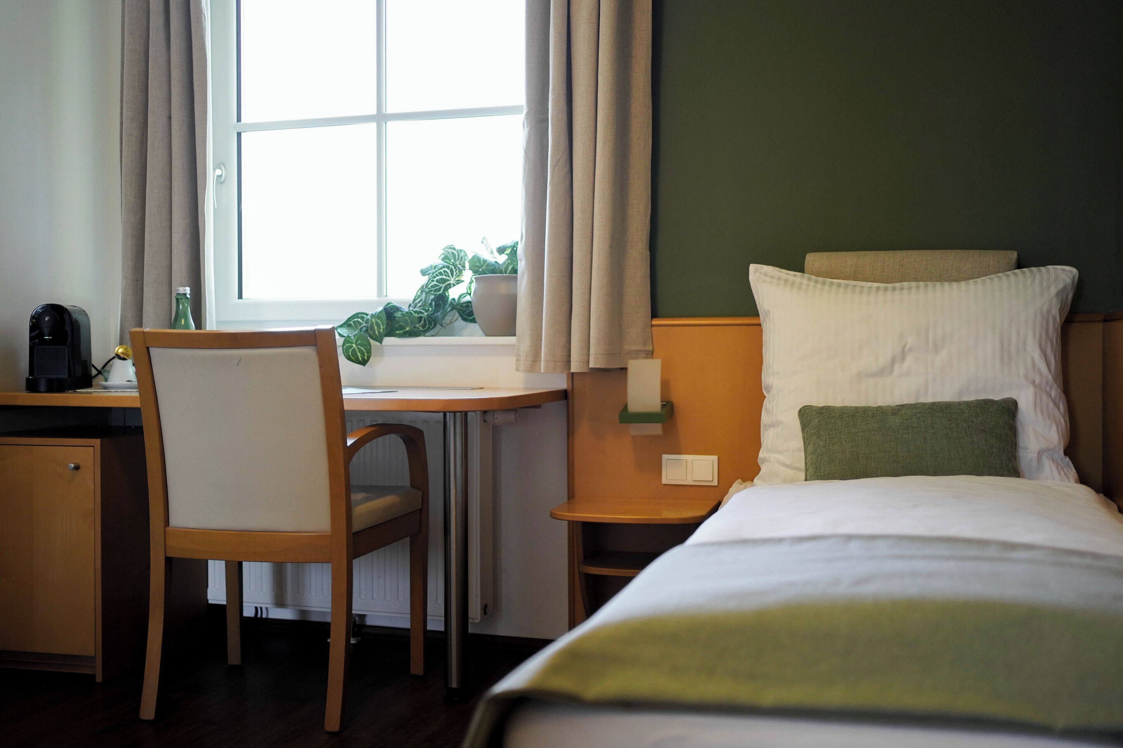 A hotel room with a bed, a desk, a chair, a small table, a window with curtains, and a plant.