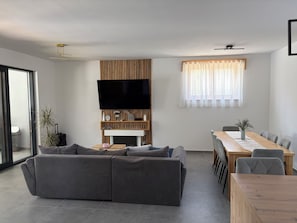A modern living room with a sectional sofa, a dining table with chairs, a flat-screen TV mounted on a wooden panel, and a large window with sheer curtains.