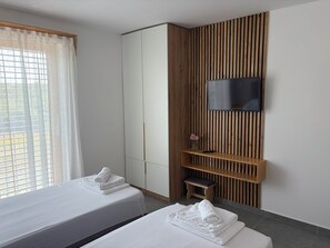 Ein Hotelzimmer mit zwei Betten, einem an einer Holzpaneele Wand montierten Fernseher und einem mit Vorhängen versehenen Fenster.