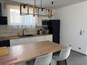 A modern kitchen with a wooden table, two chairs, a black refrigerator, a coffee machine, and a window with sheer curtains. - Modern villa with pool, jacuzzi, and sauna for relaxation and family getaway. (Maglenča)