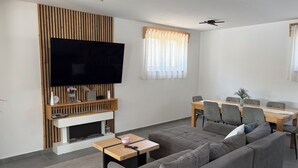 Una sala de estar moderna con una televisión de pantalla plana, chimenea, comedor con mesa y sillas, y un sofá grande de color gris.