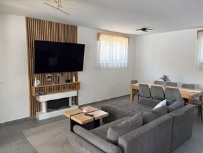 Una sala de estar moderna con una televisión de pantalla plana, chimenea, comedor con mesa y sillas, y un sofá grande de color gris.