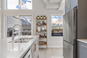 Une cuisine moderne avec un réfrigérateur en acier inoxydable, une étagère en bois ornée d’objets décoratifs et une vue sur les montagnes, visible à travers de grandes fenêtres.