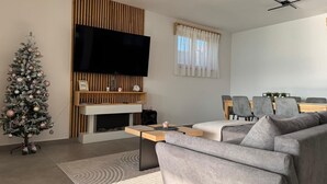 Una sala de estar moderna con una televisión de pantalla plana, un árbol de Navidad, una chimenea y un comedor con sillas grises.