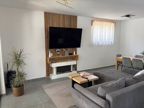 A modern living room with a flat-screen TV mounted on a wooden panel, a sofa, a coffee table, and a dining area with chairs. - Modern villa with pool, jacuzzi, and sauna in Bilogora for relaxing getaway. (Maglenča)
