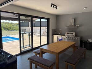 A modern kitchen with a built-in oven, a dining table with benches, and a sliding glass door leading to an outdoor area with a pool. - Modern villa with pool, jacuzzi, and sauna in Bilogora for relaxing getaway. (Maglenča)