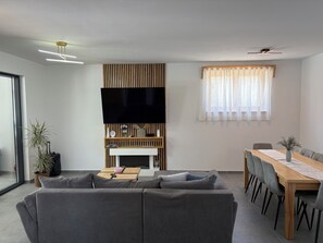 Un salon moderne comprenant un canapé, un téléviseur à écran plat, un meuble TV en bois, une table à manger avec des chaises et une fenêtre agrémentée de rideaux légers.