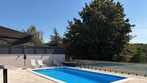 Ein rechteckiger Swimmingpool mit gefliestem Sonnendeck, zwei weißen Liegestühlen und einem schwarzen Sonnenschirm.
