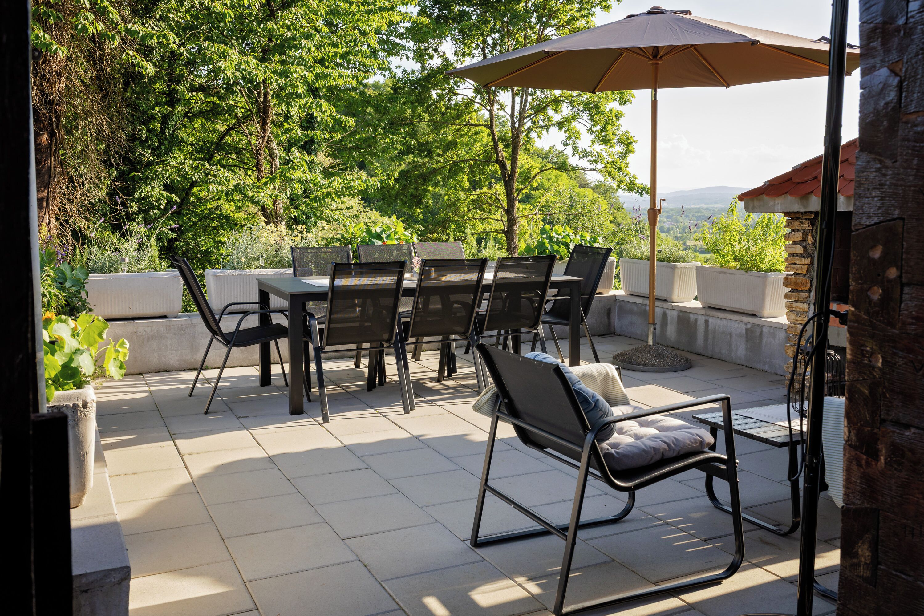 En udendørs terrasse med et bord og stole, en stor parasol og udsigt til træer og bakker.