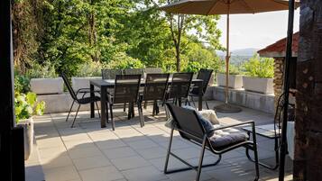 En udendørs terrasse med et bord og stole, en stor parasol og udsigt til træer og bakker.