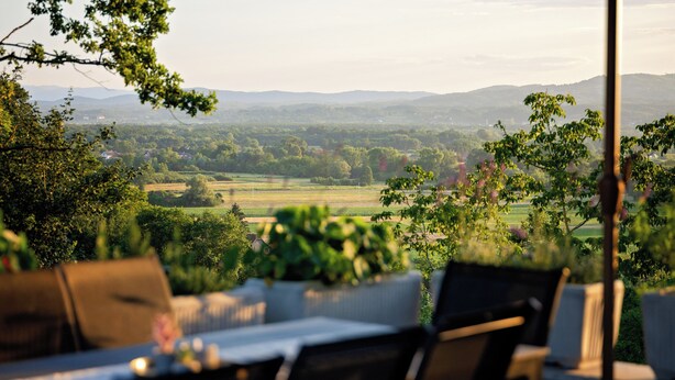 Eine Terrasse mit Sitzgelegenheiten, Blick auf eine grüne Landschaft und ferne Hügel.