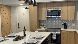 A modern kitchen with wooden cabinets, a stainless steel microwave, and a central island with a wine bottle and glasses. - Sunrise Ascent - Lock-Off - Pool & Hot Tub (Canmore)