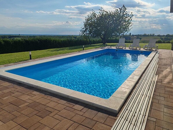 A rectangular swimming pool with a clear blue water surface, surrounded by a paved deck and a grassy area with a tree.