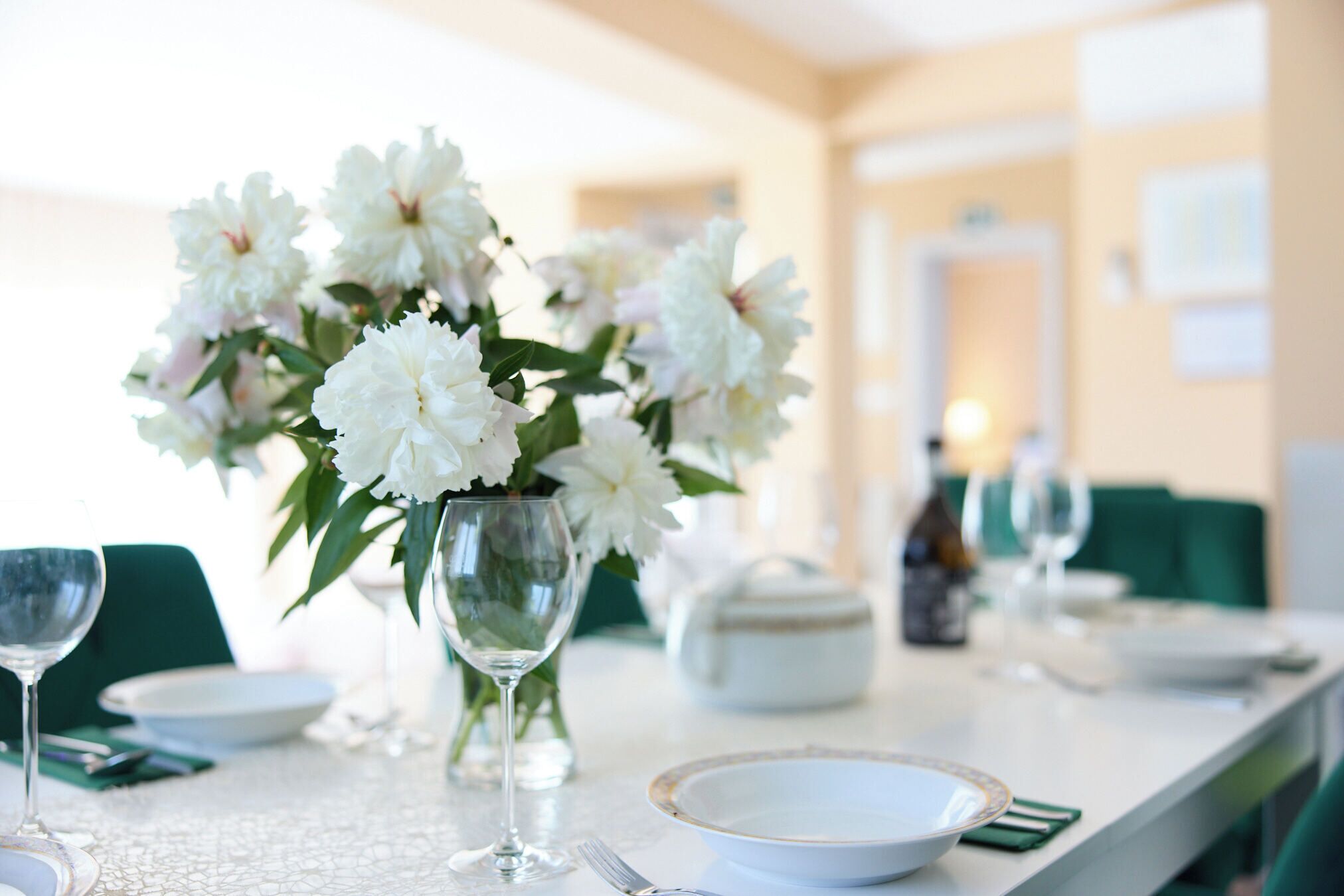 Une table à manger dressée avec de l’assiette blanche, des verres à vin transparents et un vase rempli de fleurs blanches.