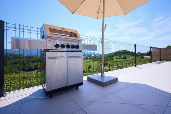 A stainless steel outdoor grill with four burners, a control panel, and a built-in oven, positioned on a patio with a view of a green landscape.