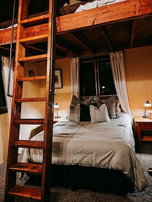 A bedroom with a wooden ladder, a bed with pillows, a nightstand with a lamp, and a window with curtains.