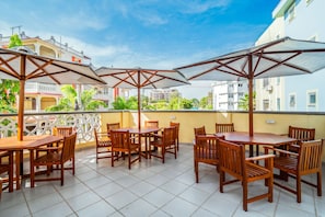 A terrace with wooden tables and chairs, umbrellas, and a view of buildings and greenery. -  2-bedroom apartment  in Nyali/AC, 2 mins walk to the beach with Gym and pool. (Mombasa)
