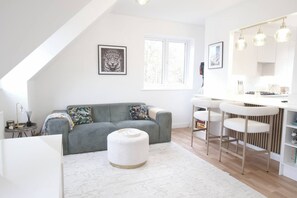 A modern living room with a grey sofa, a white ottoman, and a kitchen area with bar stools and a white countertop. - The Lily  Suite with parking (Berkshire)