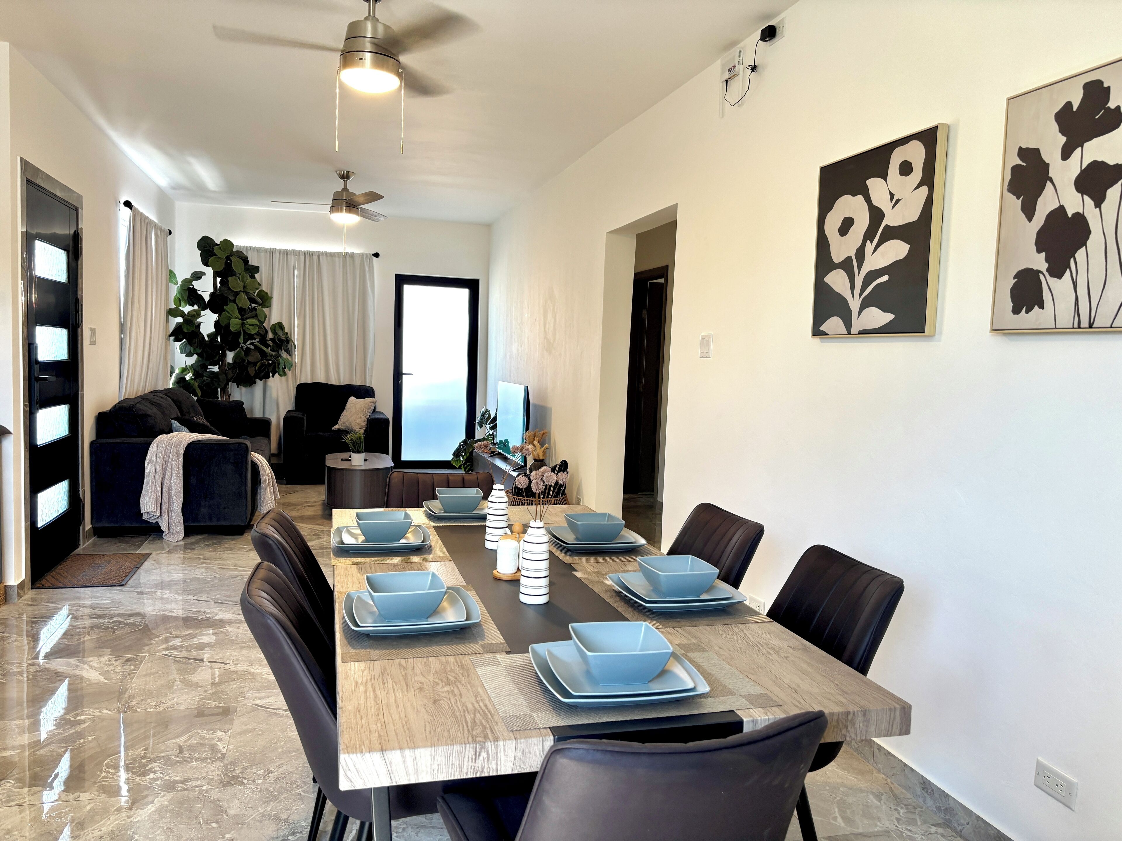 A dining area with a wooden table set for six, surrounded by black chairs. A large potted plant is in the background, and there are two ceiling fans.