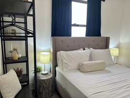 Una cama bien hecha con sábanas y almohadas blancas, una lámpara de noche, una mesita de madera pequeña con una lámpara y un florero, y una estantería metálica negra con objetos decorativos.