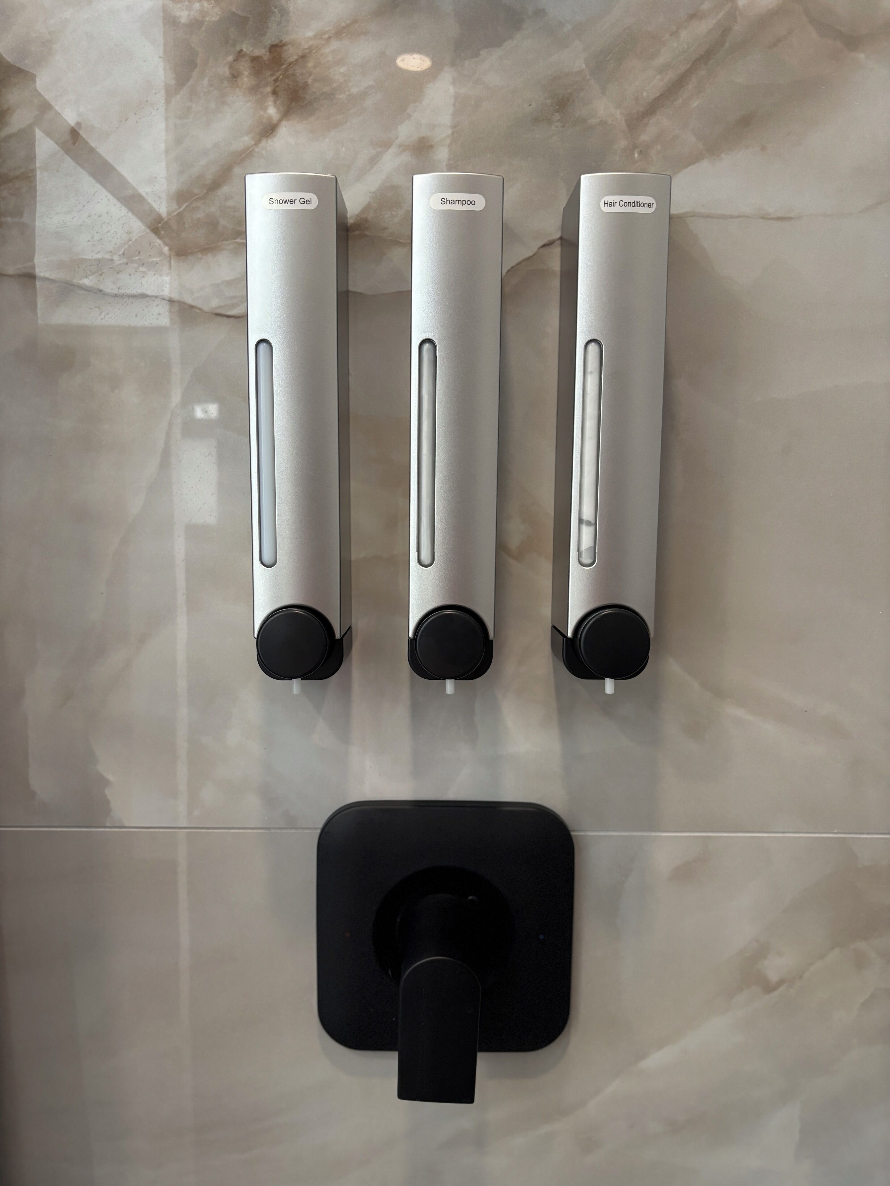 Dispensador montado en la pared con tres compartimentos etiquetados para gel de ducha, champú y acondicionador para cabello.