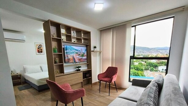 A modern living room with a sofa, two chairs, a TV, and a large window with a city view. - Cali - Edificio Miro Living Refugio (Cali)