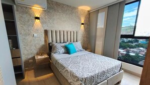 A bedroom with a large bed, two bedside tables, a wardrobe, and a view of the city through a large window. - Cali - Edificio Miro Living Refugio (Cali)