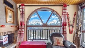 A cozy room with a fireplace, a leather chair with a deer pillow, a red table, and a view of snow-capped mountains through a large arched window.