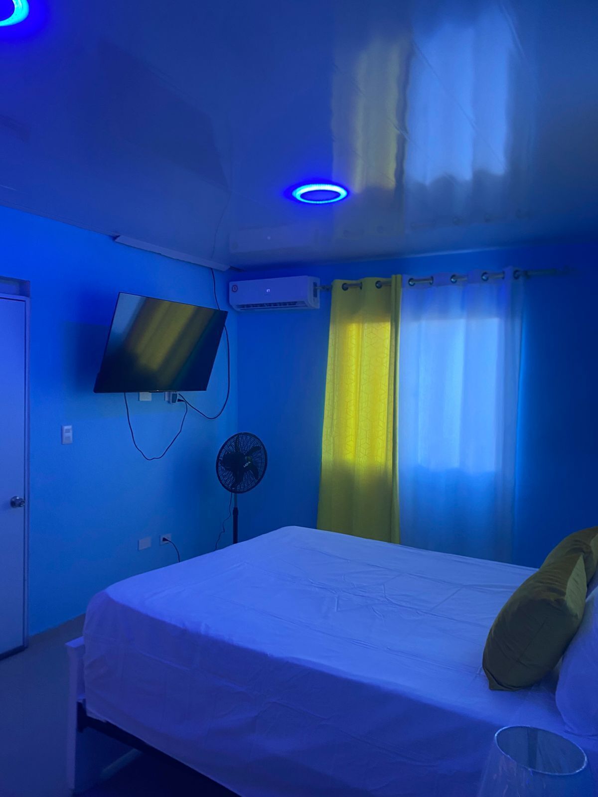 Een kamer met een bed, een aan de muur bevestigde televisie, een airconditioningapparaat, een ventilator en gordijnen.