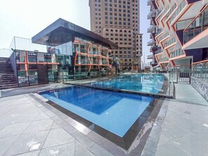 A modern outdoor swimming pool with glass railings, surrounded by a tiled deck and adjacent buildings. - Stayshort - Magnificent 3 Bedroom Apartment in Al Jaddaf That Sleeps 8 (Dubai)