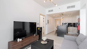 A modern living room with a flat-screen TV, a coffee table, and a grey sofa. A kitchen is visible in the background with a black countertop and stainless steel appliances. - Stayshort - Magnificent 3 Bedroom Apartment in Al Jaddaf That Sleeps 8 (Dubai)