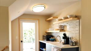 Een moderne keuken met een inbouwoven, magnetron en spoelbak. Er staat een afvalbak en een deur naar een lichte kamer met uitzicht op groen.