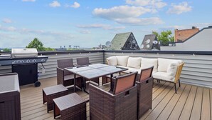 A rooftop patio with a grill, wicker furniture, and a view of the cityscape.