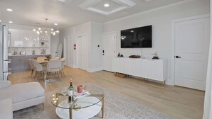 A modern living room with a dining area, white cabinetry, a flat-screen TV, and a ceiling with recessed lighting.