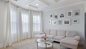 A living room with a sofa, a glass coffee table, and a rug. There are curtains on the windows, and pictures on the wall.