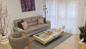 Uma sala de estar moderna com um sofá bege, uma mesa de centro de vidro e uma poltrona cinza.