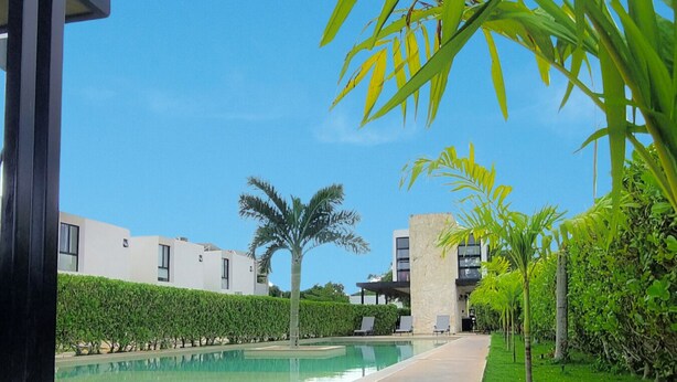 A pool area with a clear sky, palm trees, and modern buildings in the background. - Eco Loft : Balcony with bathtub : Swimming pool and Gym : North Merida (Cholul)
