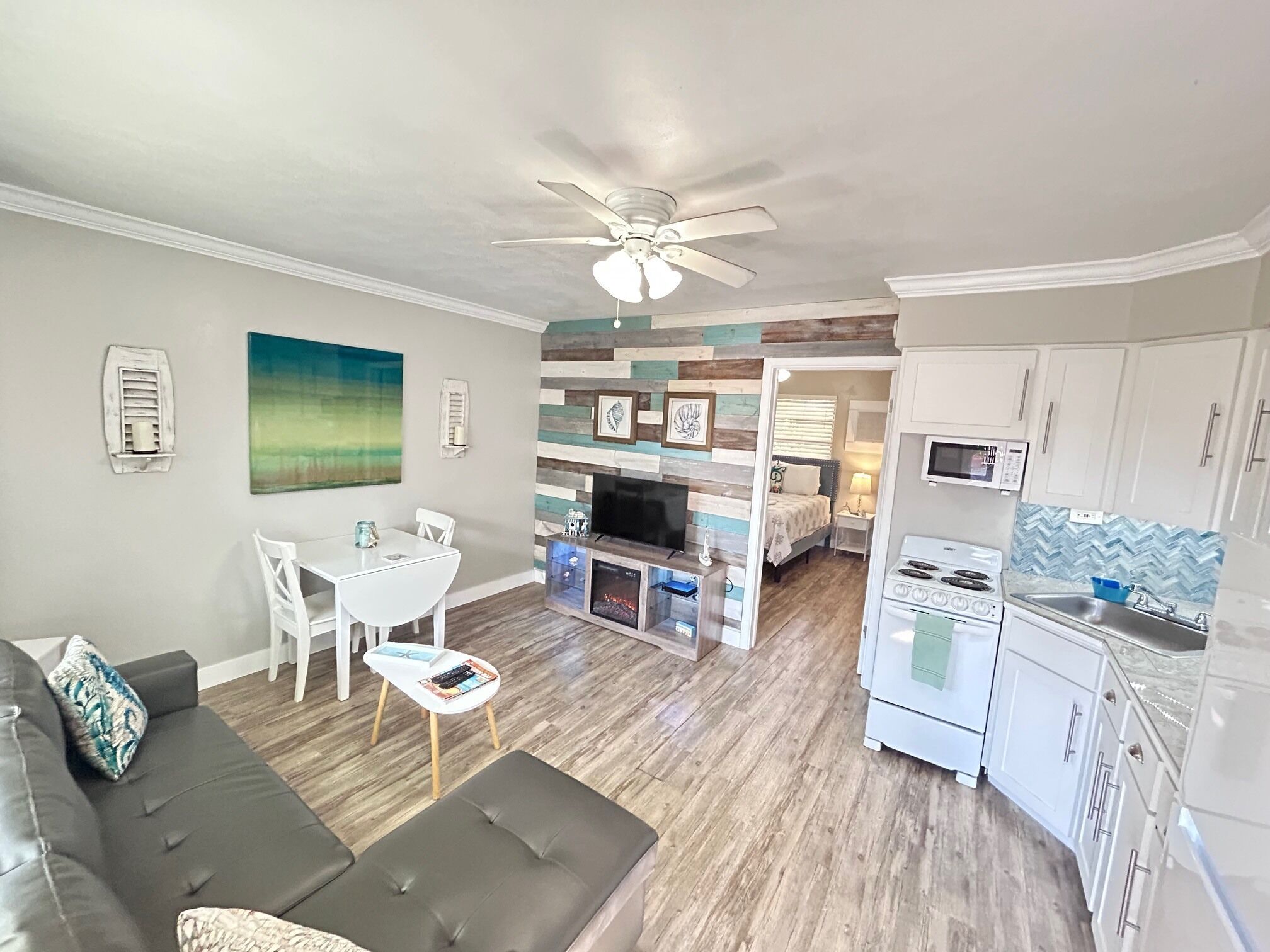 A modern living room with a gray sofa, a dining area with a white table and chairs, a kitchen with white cabinets and appliances, and a ceiling fan.
